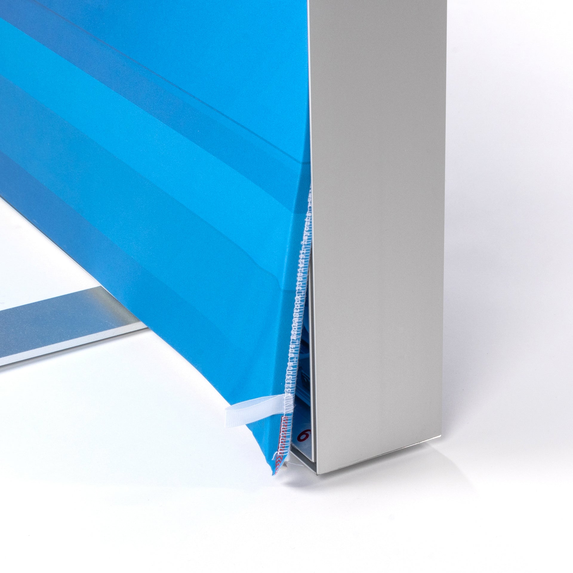 Sichtbar ist die untere Ecke eines Aluminiumrahmens, Blau bedrucktes Textil ist teilweise aus dem Rahmen gelöst, Am Stoffrand ist eine angenähte Kederlippe zu erkennen, Kleine weiße Zuglasche erleichtert das Herausziehen des Textils, Rechts ist das glatte, silberfarbene Rahmenprofil zu sehen, Bild zeigt, wie das Textil in den Rahmen eingesetzt oder gewechselt wird