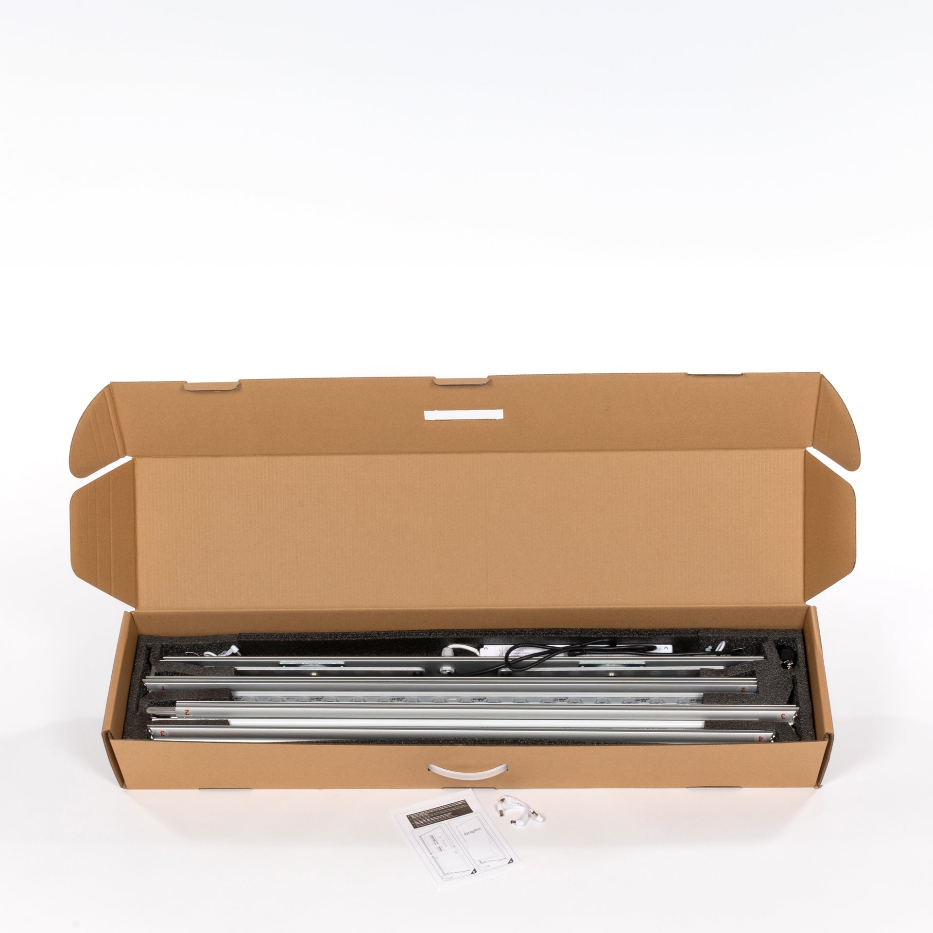 Geöffneter Versandkarton aus brauner Wellpappe, Innen liegen mehrere silberfarbene Aluminiumprofile, Profile sind mit schwarzem Schaumstoff geschützt, Sichtbare Kabel und technische Bauteile im Karton, Vor dem Karton liegen Anleitung und kleines Kabel, Verpackung zeigt den Lieferumfang vor der Montage