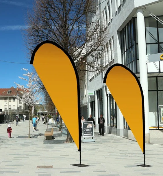 Beachflag Drop Austauschdruck: Zwei tropfenförmige, orangefarbene Beachflags stehen auf einem grauen Gehweg. Die Flags sind an schwarzen Masten befestigt, die wiederum auf schwarzen, rechteckigen Standfüßen stehen, in Wiesbaden bei City Medien Einkaufswelt für Partner