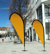 Beachflag Drop Austauschdruck: Zwei tropfenförmige, orangefarbene Beachflags stehen auf einem grauen Gehweg. Die Flags sind an schwarzen Masten befestigt, die wiederum auf schwarzen, rechteckigen Standfüßen stehen, in Wiesbaden bei City Medien Einkaufswelt für Partner