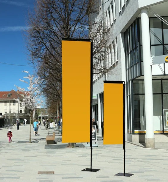 Beachflag Rechteck Medium 290 cm: Zwei rechteckige, orangefarbene Beachflags in unterschiedlicher Höhe. Sie sind einfarbig, an schwarzen Masten befestigt und stehen auf schwarzen, quadratischen Standfüßen auf einem grauen Gehweg, in Wiesbaden bei City Medien Einkaufswelt für Partner