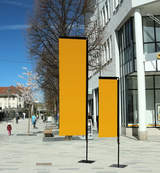 Beachflag Rechteck Large 400 cm: Zwei rechteckige, orangefarbene Beachflags in unterschiedlicher Höhe. Sie sind einfarbig, an schwarzen Masten befestigt und stehen auf schwarzen, quadratischen Standfüßen auf einem grauen Gehweg, in Wiesbaden bei City Medien Einkaufswelt für Partner