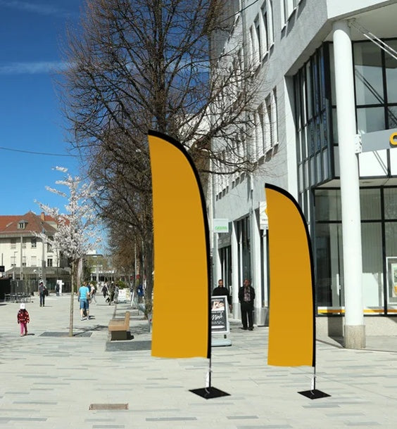 Beachflag Wind Extra Small 215 cm: Zwei windförmige, orangefarbene Beachflags in unterschiedlicher Größe stehen auf einem grauen Gehweg. Die Flags sind an schwarzen Masten befestigt, die auf schwarzen, quadratischen Standfüßen stehen, in Wiesbaden bei City Medien Einkaufswelt für Partner