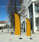 Beachflag Wind Medium 320 cm: Zwei windförmige, orangefarbene Beachflags in unterschiedlicher Größe stehen auf einem grauen Gehweg. Die Flags sind an schwarzen Masten befestigt, die auf schwarzen, quadratischen Standfüßen stehen, in Wiesbaden bei City Medien Einkaufswelt für Partner