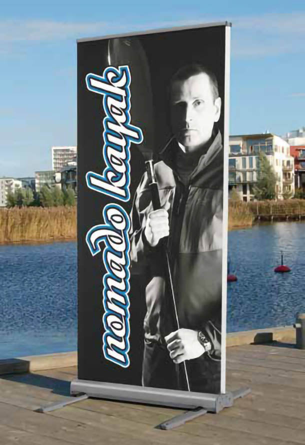 Roll Up Outdoor B 85 cm x H 200 cm: Ein aufgestelltes Roll-Up-Display im Freien. Das Display ist mit einem schwarz-weißen Foto eines Mannes mit einem Paddel und dem Schriftzug „nomad kayaker“ bedruckt. Der Ständer hat an den Seiten vier ausklappbare Füße, in Wiesbaden bei City Medien Einkaufswelt für Partner
