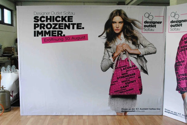 Textilfaltwand: Eine gerade Textilfaltwand mit einem Werbedruck für das „Designer Outlet Soltau“. Der Druck zeigt eine Frau mit einer pinken Handtasche und dem Text „SCHICKE PROZENTE. IMMER.“, in Wiesbaden bei City Medien Einkaufswelt für Partner