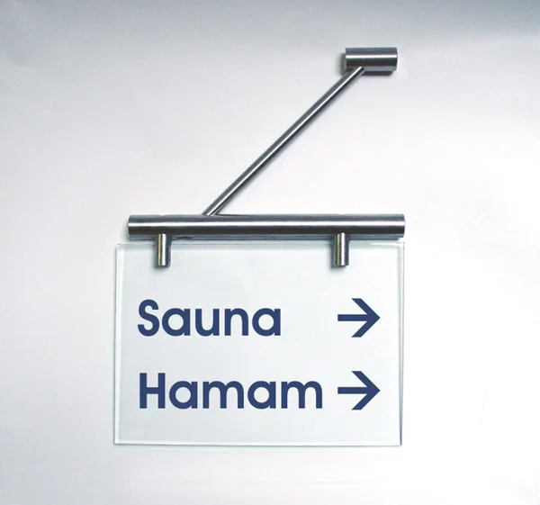 Fahnenschild System Seeshaupt: Ein rechteckiges, transparentes Schild mit silbernem Halter. Darauf ist in blau die Wörter Sauna und Hamam abgebildet. Das Schild ist quer an einer silbernen Halterung an der Wand montiert, in Wiesbaden bei City Medien Einkaufswelt für Partner