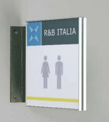 Fahnenschild System Seeshaupt: Ein quadratisches, zweifarbiges Fahnenschild. Der obere Bereich ist dunkelgrau mit dem weißen Schriftzug „R&B ITALIA“ und einem blauen Symbol. Der untere Bereich ist weiß mit hellgrauen Piktogrammen für Damen und Herren und einem gelben Streifen. Das Schild ist an einer schwarzen Halterung an der Wand montiert, in Wiesbaden bei City Medien Einkaufswelt für Partner