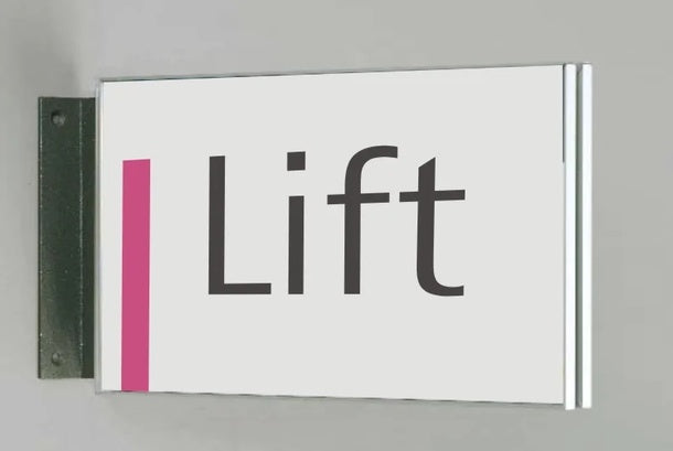 Fahnenschild System Seeshaupt: Ein querformatiges, weißes Schild mit silbernem Rahmen. An der linken Seite befindet sich ein vertikaler, rosafarbener Streifen. In der Mitte steht das Wort „Lift“ in grauer Schrift. Das Schild ist an einer schwarzen Halterung an der Wand montiert, in Wiesbaden bei City Medien Einkaufswelt für Partner