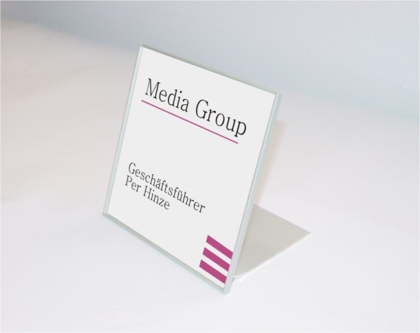 Tischaufsteller System Karlsruhe: Ein silberner Tischaufsteller mit einem quadratischen bedruckten Blatt. Der Druck ist weiß und hat den schwarzen Schriftzug „Media Group“, „Geschäftsführer Per Hinze“ und ein lilafarbenes Logo am unteren Rand, in Wiesbaden bei City Medien Einkaufswelt für Partner