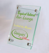 Tischaufsteller System Seeshaupt: Ein rechteckiger Tischaufsteller aus zwei klaren Acrylglasscheiben. Der Druck zwischen den Scheiben zeigt die Aufschrift „Tropical Island Bar-Lounge“ in grüner und „Daiquiri“ in grüner und roter Schrift. In der Mitte steht der gelbe Schriftzug „Cocktail des Monats“. Der Aufsteller wird von vier silbernen Metallstiften zusammengehalten, in Wiesbaden bei City Medien Einkaufswelt für Partner