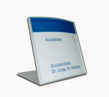 Tischaufsteller System Zenit: Ein silberner Tischaufsteller mit einem geschwungenen oberen Rand. Er zeigt ein bedrucktes Papier mit einem blauen Streifen am oberen Rand und der blauen Aufschrift „Assistenten“ sowie „Assistenzärzte Dr. Lange, Dr. Eichhorn“, in Wiesbaden bei City Medien Einkaufswelt für Partner