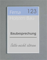 Wandschild System Karlsruhe: Ein rechteckiges Schild mit einem silbernen Rahmen. Es ist dreigeteilt in einen hellblauen Bereich mit der Aufschrift „Firma 123“, einen hellgrauen Bereich mit „Holsten Bau“ und einen weißen Bereich mit dem Text „Baubesprechung“ und „Bitte nicht stören“. Das Schild ist an einer grauen Wand befestigt, in Wiesbaden bei City Medien Einkaufswelt für Partner