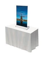 Roll Up Mini Deluxe DIN A4: Ein bedrucktes Roll-Up-Display im DIN A4 Format. Das Display zeigt eine Windmühle an einem Fluss und steht auf einer weißen, mit einem Tuch bedeckten Theke, in Wiesbaden bei City Medien Einkaufswelt für Partner
