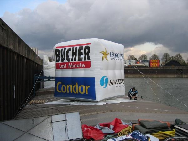 Aufblasbare Sonderproduktionen: Ein großer, aufblasbarer, würfelförmiger Werbeartikel, der mit den Logos von „BUCHER“, „Condor“, „IBEROSTAR“ und „SENTIDO“ bedruckt ist. Er steht auf einer Metallplattform an einem Ufer, in Wiesbaden bei City Medien Einkaufswelt für Partner