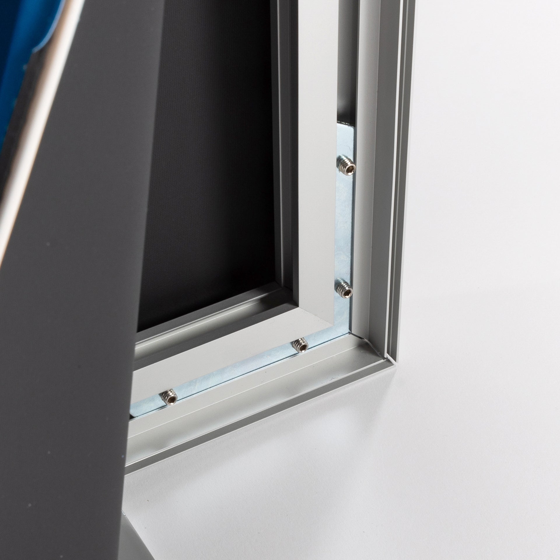 Detailaufnahme einer Rahmen-Ecke, Silberfarbenes Aluminiumprofil mit innerer Nut, Schwarze Textilrückseite teilweise nach vorne geklappt, Eckverbindung mit Metallplatte sichtbar, Mehrere Schrauben und Gewindebolzen zu erkennen, Weiße und graue Rahmenteile liegen sauber ineinander, Bild zeigt die stabile Innenkonstruktion des Textil-Spannrahmens