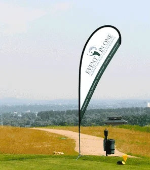 Beachflag Drop Small 210 cm: Eine tropfenförmige Beachflag mit dem weiß-grünen Logo „EVENT IN ONE“ steht auf einer Wiese neben einem Sandweg. Die Flagge ist auf einem schwarzen Mast befestigt, in Wiesbaden bei City Medien Einkaufswelt für Partner