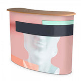 Messetheke Pop-Up 2x2: Eine geschwungene Messetheke mit einem abstrakten, bedruckten Stoff in den Farben Pastellrosa, Mintgrün, Dunkelgrau und einem weißen Motiv eines weiblichen Kopfs. Die Tischplatte ist aus hellbraunem Holz, in Wiesbaden bei City Medien Einkaufswelt für Partner