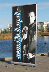 Roll Up Outdoor B 85 cm x H 200 cm: Ein aufgestelltes Roll-Up-Display im Freien. Das Display ist mit einem schwarz-weißen Foto eines Mannes mit einem Paddel und dem Schriftzug „nomad kayaker“ bedruckt. Der Ständer hat an den Seiten vier ausklappbare Füße, in Wiesbaden bei City Medien Einkaufswelt für Partner