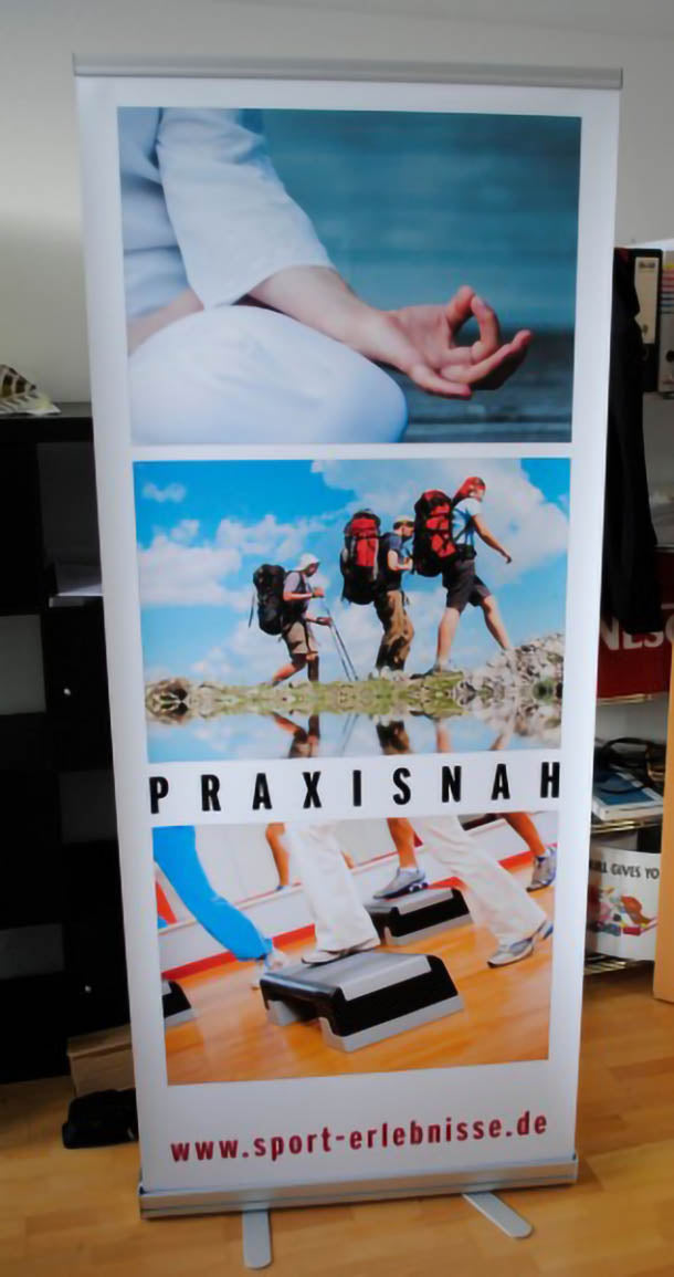 Roll Up Standard B 150 cm x H 200 cm: Ein aufgestelltes Roll-Up-Display mit einem bedruckten Banner. Das Banner zeigt drei Bilder, die von oben nach unten eine Hand in der Meditation, zwei Wanderer und eine Step-Aerobic-Gruppe zeigen. Unter dem mittleren Bild steht „PRAXISNAH“ und unten die URL „www.sport-erlebnisse.de“, in Wiesbaden bei City Medien Einkaufswelt für Partner