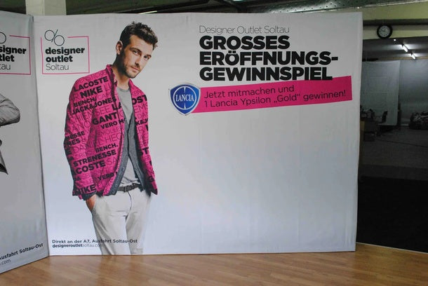 Textilfaltwand: Eine gerade Textilfaltwand mit einem Werbedruck für das „Designer Outlet Soltau“. Der Druck zeigt einen Mann in einem pinken Anzug, in Wiesbaden bei City Medien Einkaufswelt für Partner