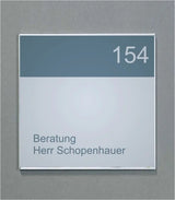Wandschild System Karlsruhe: Ein quadratisches Schild mit einem silbernen Rahmen. Der obere Bereich ist dunkelgrau mit der weißen Zahl „154“. Der untere Bereich ist hellgrau mit dem grauen Text „Beratung Herr Schopenhauer“. Das Schild ist an einer grauen Wand befestigt, in Wiesbaden bei City Medien Einkaufswelt für Partner