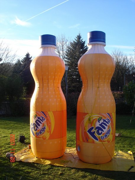 Aufblasbare Sonderproduktionen: Zwei aufblasbare, orangefarbene Fanta-Flaschen stehen auf einem gelben Tuch in einem Garten. Die Flaschen sind originalgetreu bedruckt und haben blaue Schraubverschlüsse, in Wiesbaden bei City Medien Einkaufswelt für Partner