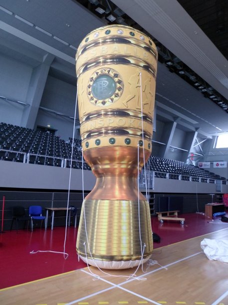 Aufblasbare Sonderproduktionen: Ein großer, aufblasbarer DFB-Pokal steht in einer Sporthalle. Er ist goldfarben mit grünen, roten und blauen Details. Weiße Seile halten ihn am Boden, in Wiesbaden bei City Medien Einkaufswelt für Partner