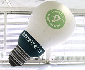 Aufblasbare Sonderproduktionen: Eine aufblasbare, weiße Glühbirne hängt von einer Decke. Sie hat ein grünes, rundes Logo mit dem Text „SAUBERES LICHT SAUBER RECYCELT“ und darunter „lichtzeichen.de“ auf einem schwarzen Streifen, in Wiesbaden bei City Medien Einkaufswelt für Partner